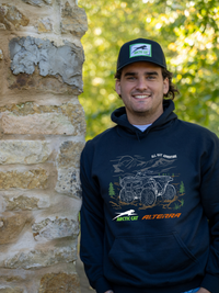 A smiling man wearing a black hoodie and matching black cap stands next to a rustic stone wall with a blurred green forest background. The hoodie features a graphic of an ATV with the text &