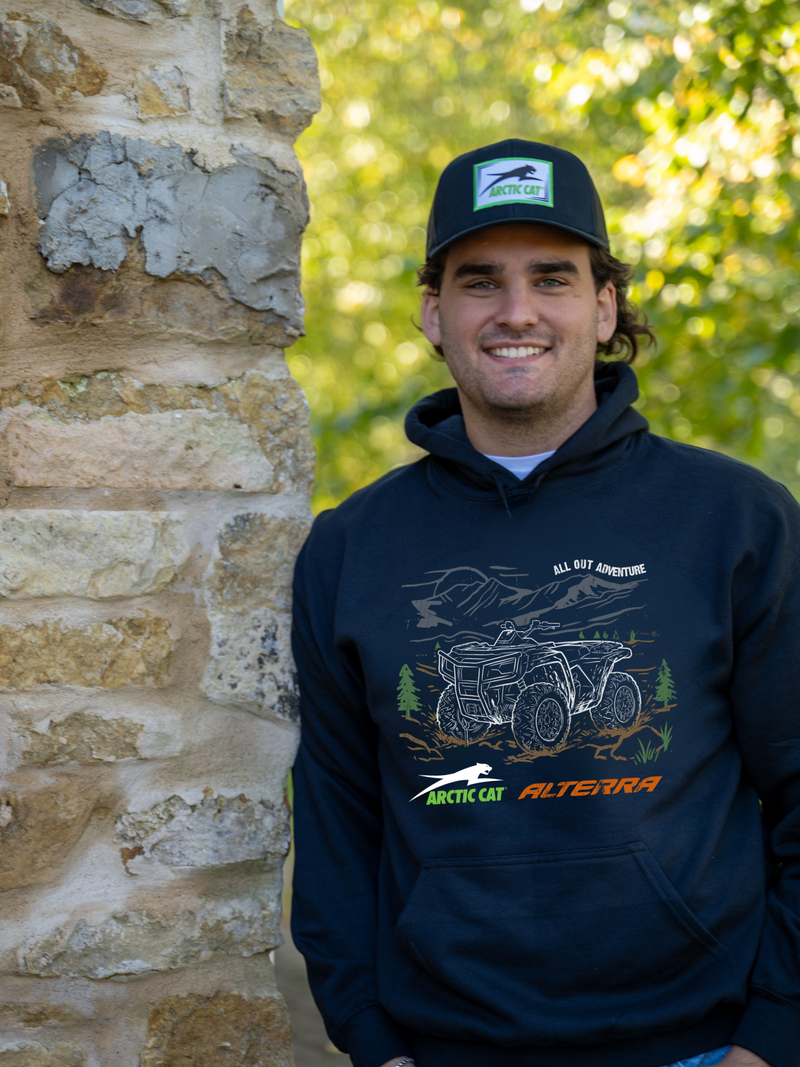 A smiling man wearing a black hoodie and matching black cap stands next to a rustic stone wall with a blurred green forest background. The hoodie features a graphic of an ATV with the text &