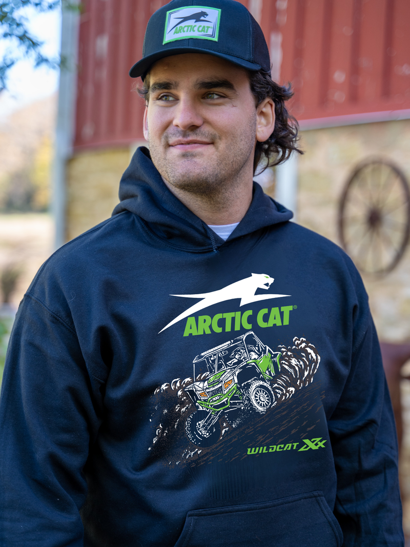 A man wearing a navy blue Arctic Cat hoodie with a green and white graphic of a dune buggy, standing in front of a rustic red barn background.