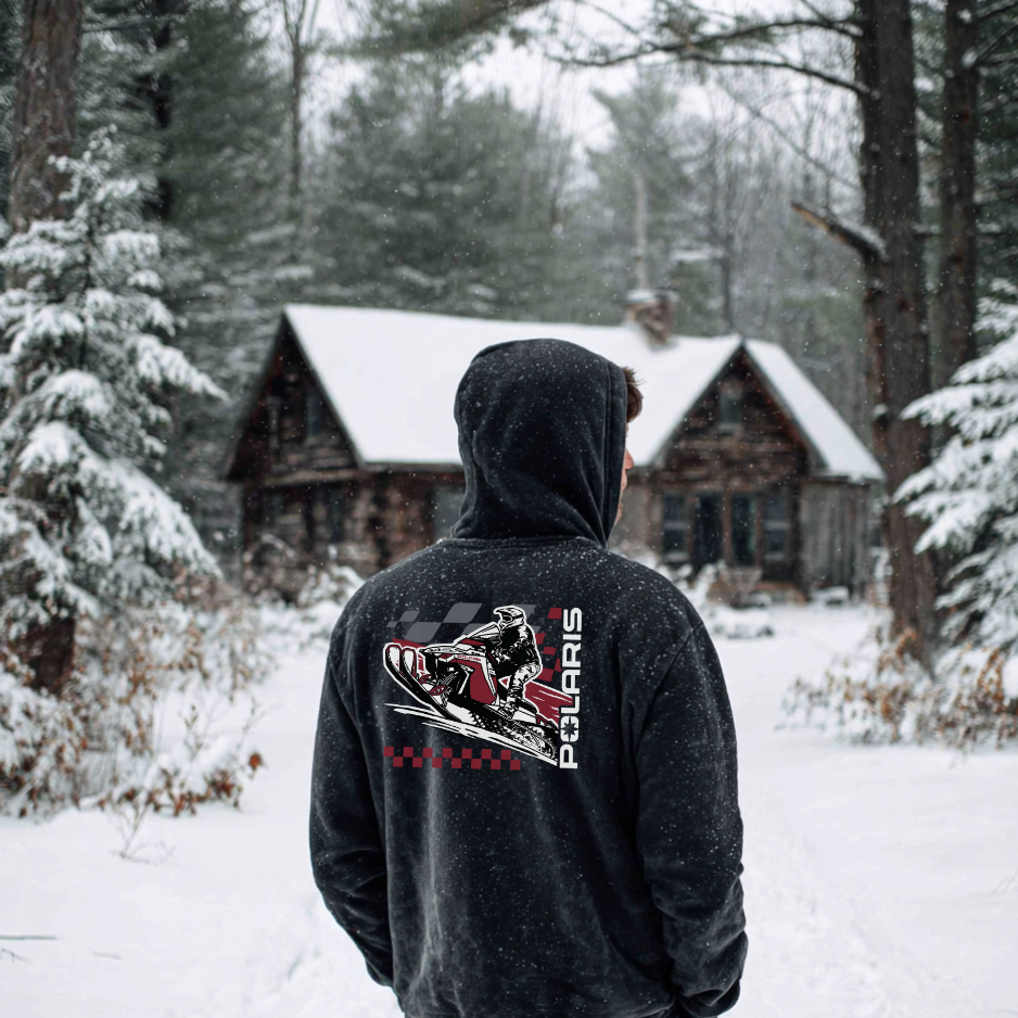 Person wearing a hoodie with Polaris logo in a snowy forest with log cabin.