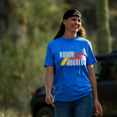 Woman wearing a blue 'Rough & Rugged' t-shirt outdoors with a blurred background