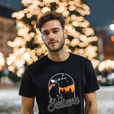 Man wearing a black t-shirt with a graphic design and the word 'Believe' in front of blurred lights.