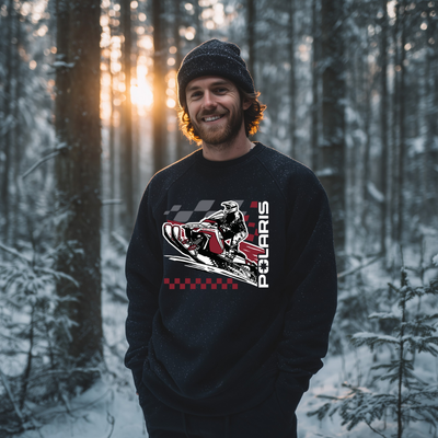 Man wearing a black Polaris sweatshirt in a snowy forest with sunlit trees.